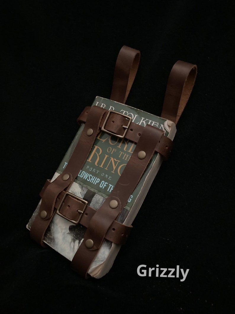May include: A brown leather book carrier with buckles and straps, holding a copy of "The Lord of the Rings: The Fellowship of the Ring." The carrier has two loops for carrying. The word "Grizzly" is in the bottom right corner.