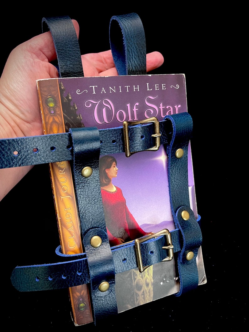 May include: A navy blue leather book holder with brass buckles and rivets, holding a book titled "Wolf Star" by Tanith Lee. The holder has straps for carrying and securing the book. The book cover features a woman in a red dress.