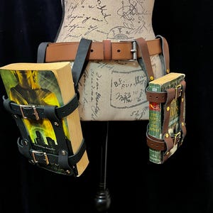May include: A brown leather belt with two book holders. The holders are black leather with silver buckles. Each holder contains a book. The belt is displayed on a mannequin.