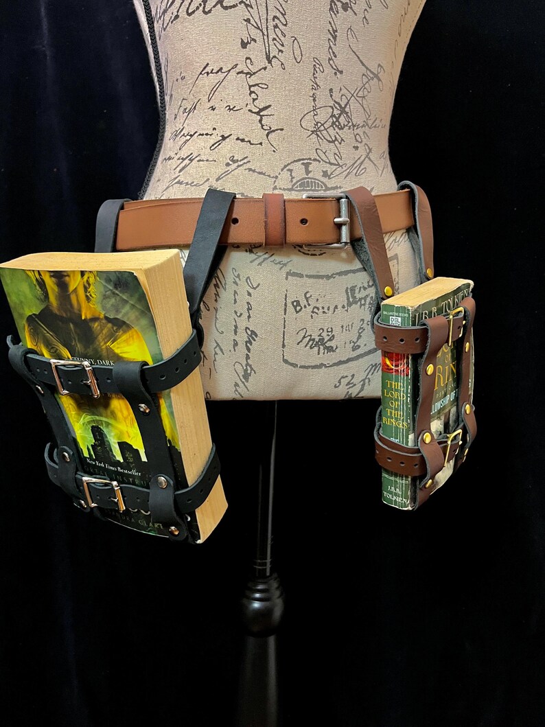 May include: A brown leather belt with two book holders. The holders are black leather with silver buckles. Each holder contains a book. The belt is displayed on a mannequin.