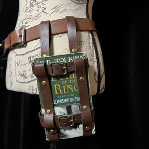 May include: Brown leather belt with a book holder. The book holder is attached to the belt with straps and buckles. The book is titled "The Lord of the Rings: Part One: Fellowship of the Ring".