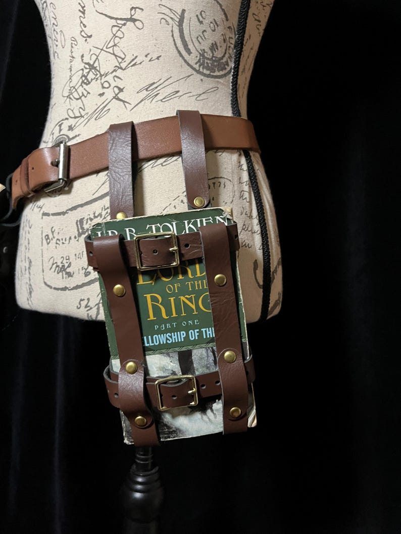 May include: Brown leather belt with a book holder. The book holder is attached to the belt with straps and buckles. The book is titled "The Lord of the Rings: Part One: Fellowship of the Ring".