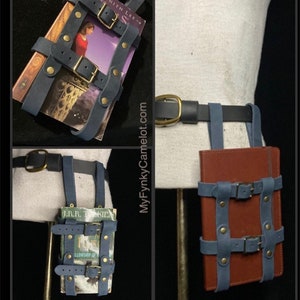 May include: A blue leather book holder with brass buckles and rivets, designed to be worn on a belt. The holder is shown with a book inside. The belt is black with a gold buckle. The website MyFynkyCamelot.com is visible.