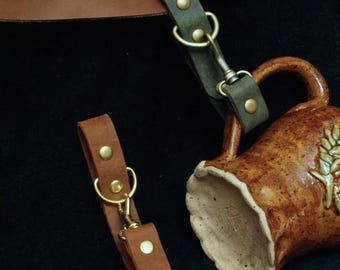 Könnte beinhalten: Ein brauner Bierkrug aus Keramik mit Henkel und Weizenmuster, begleitet von Lederriemen und Metallverschlüssen. Die Riemen sind braun und olivgrün, mit messingfarbener Hardware. Der Krug hat eine strukturierte, gesprenkelte Oberfläche.