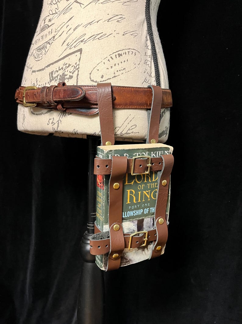 May include: A brown leather book holder with brass buckles and straps, designed to be worn on a belt. The book holder is attached to a brown leather belt. The book inside is titled "LORD OF THE RING" and "FELLOWSHIP OF THE".