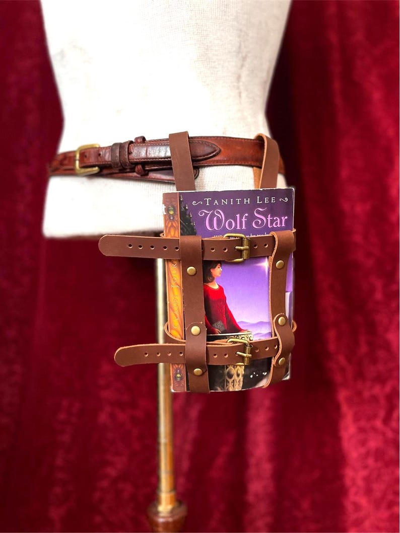 May include: A brown leather book holder attached to a brown leather belt with a gold buckle. The book holder is designed to carry a book, and the cover of the book reads "Wolf Star" by Tanith Lee.