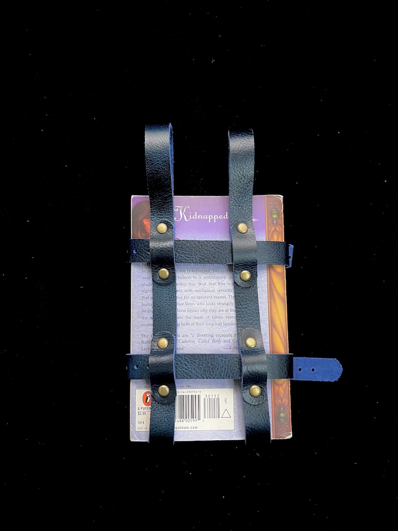 May include: Black leather book holder with brass rivets, holding a paperback book. The book has a purple cover with the title "Kidnapped". The holder has adjustable straps.