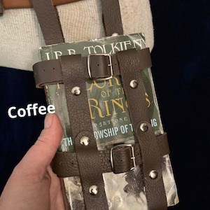 May include: A brown leather book holder with silver rivets and buckles, designed to be worn on a belt. The holder contains a book with the title "The Fellowship of the Ring" by J.R.R. Tolkien. The word "Coffee" is written on the image.
