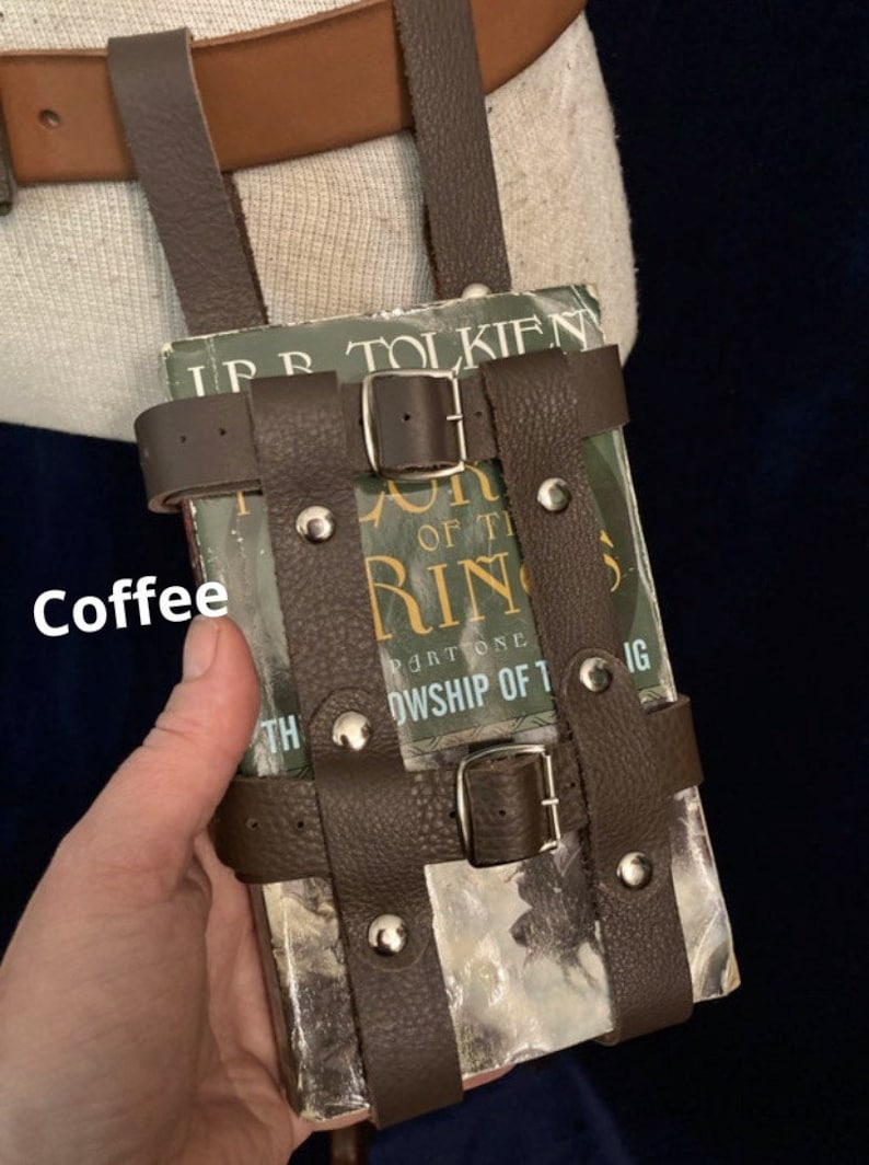 May include: A brown leather book holder with silver rivets and buckles, designed to be worn on a belt. The holder contains a book with the title "The Fellowship of the Ring" by J.R.R. Tolkien. The word "Coffee" is written on the image.