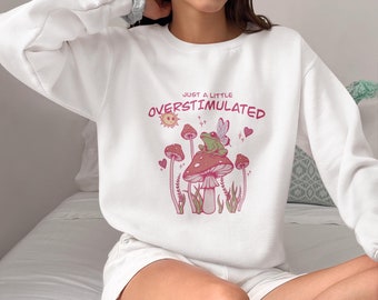 May include: White sweatshirt with a graphic of a frog wearing fairy wings sitting on a mushroom. The text "Just a little Overstimulated" is printed on the sweatshirt.
