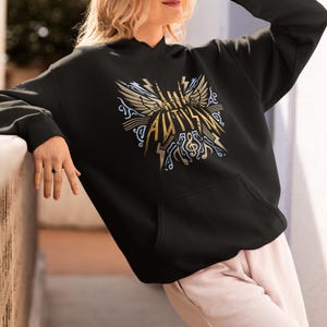 Puede incluir: Sudadera con capucha negra con un diseño gráfico dorado y azul. El diseño presenta alas, notas musicales y la palabra "AVIAN" en letras doradas. La sudadera tiene un bolsillo delantero y mangas largas.