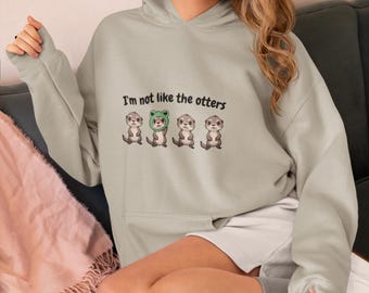 Op de afbeelding: Een lichtgrijze hoodie met een afbeelding van vier otters, waarvan er één een groene kikkerhoed draagt. De tekst "Ik ben niet zoals de anderen" staat op de hoodie gedrukt.