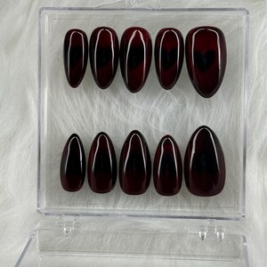 May include: A clear plastic box containing ten red and black oval-shaped false nails with a heart design.