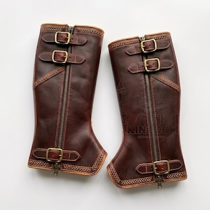 May include: A pair of brown leather riding boots with brass-colored buckles and zippers. The boots have decorative stitching and a light brown trim. The boots are knee-high and have three buckles and a zipper closure.