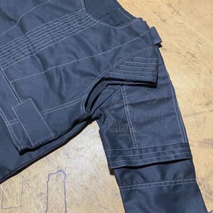 May include: Close-up of a black garment with white stitching details. The fabric appears to be a sturdy material, with visible seams and reinforced areas. The design includes a collar and sleeves.
