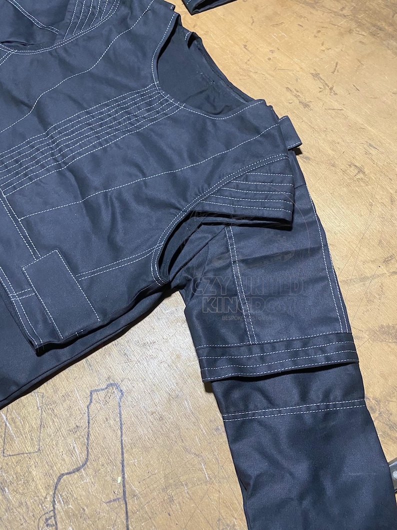 May include: Close-up of a black garment with white stitching details. The fabric appears to be a sturdy material, with visible seams and reinforced areas. The design includes a collar and sleeves.