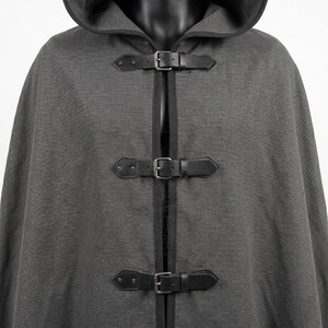 May include: A charcoal gray hooded cloak featuring a textured fabric finish and black trim along the edges. The front closure consists of three horizontal black faux leather straps with silver-toned metal buckles and rivets for secure fastening.