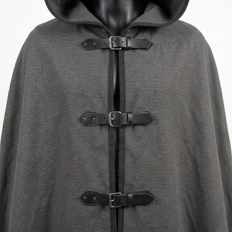 May include: A charcoal gray hooded cloak featuring a textured fabric finish and black trim along the edges. The front closure consists of three horizontal black faux leather straps with silver-toned metal buckles and rivets for secure fastening.