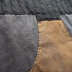 K&ouml;nnte beinhalten: Nahaufnahme eines Kleidungsst&uuml;cks, das ein Patchwork-Design zeigt. Der obere Teil ist ein dunkelgrauer, gesteppter Stoff. Darunter weist das Kleidungsst&uuml;ck eine Kombination aus grauem und braunem Stoff mit sichtbaren N&auml;hten in einem kontrastierenden gelbbraunen Faden auf.