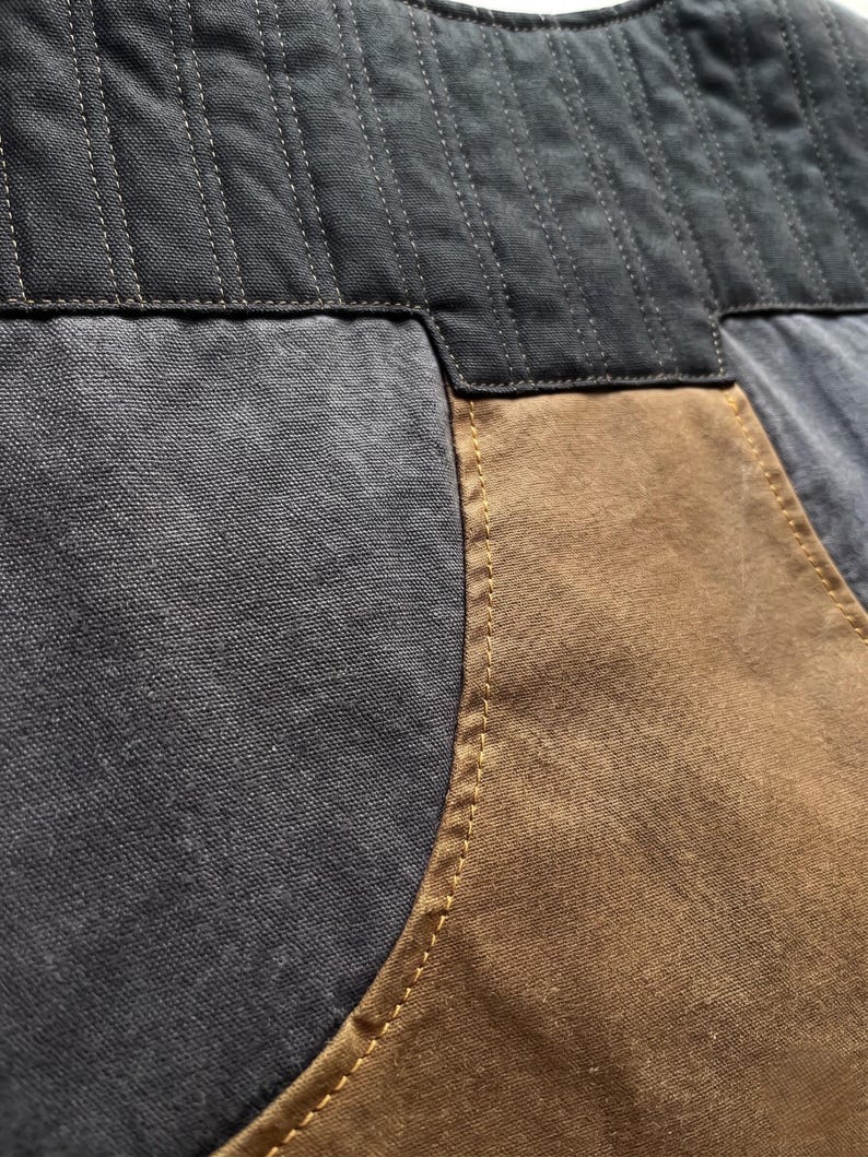 K&ouml;nnte beinhalten: Nahaufnahme eines Kleidungsst&uuml;cks, das ein Patchwork-Design zeigt. Der obere Teil ist ein dunkelgrauer, gesteppter Stoff. Darunter weist das Kleidungsst&uuml;ck eine Kombination aus grauem und braunem Stoff mit sichtbaren N&auml;hten in einem kontrastierenden gelbbraunen Faden auf.