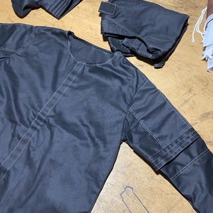 May include: A dark gray garment set, possibly outerwear, with white stitching details. The set includes a long-sleeved top and two smaller pieces, possibly pockets or other accessories. The fabric appears to be smooth and slightly shiny.