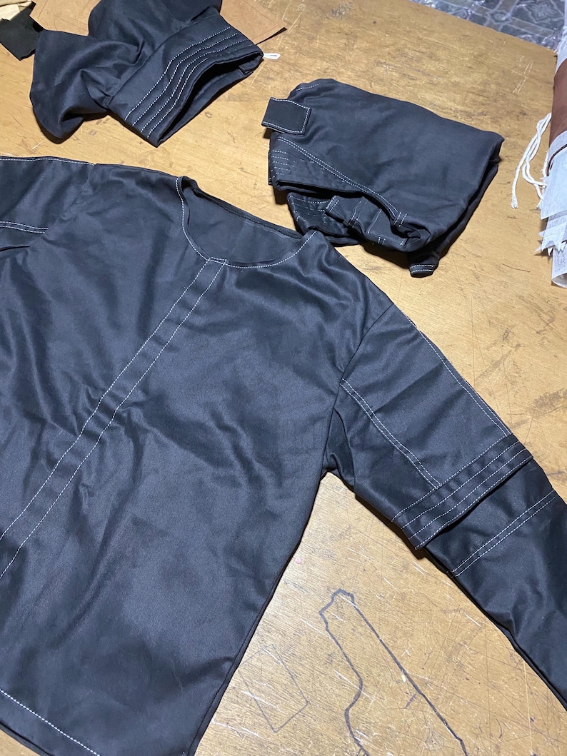 May include: A dark gray garment set, possibly outerwear, with white stitching details. The set includes a long-sleeved top and two smaller pieces, possibly pockets or other accessories. The fabric appears to be smooth and slightly shiny.