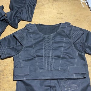 May include: Dark gray garment set with white stitching details. The set includes a short-sleeved top with a round neckline and a lower garment. The top has decorative stitching on the shoulders and front. The set is likely part of a costume.
