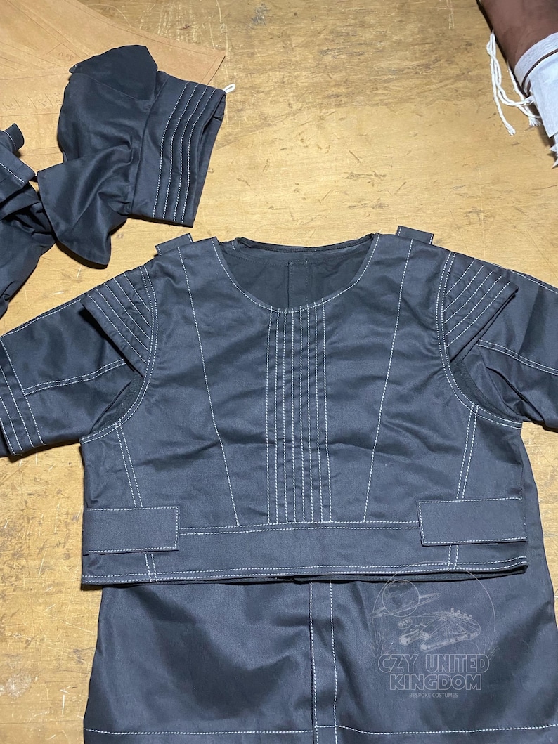 May include: Dark gray garment set with white stitching details. The set includes a short-sleeved top with a round neckline and a lower garment. The top has decorative stitching on the shoulders and front. The set is likely part of a costume.