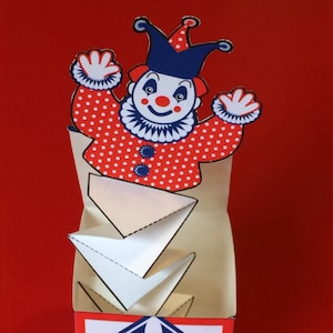 Op de afbeelding: Een rode en witte clown springt uit een doos met een blauw en wit geometrisch patroon. De clown heeft een rode neus en een blauwe en witte hoed.