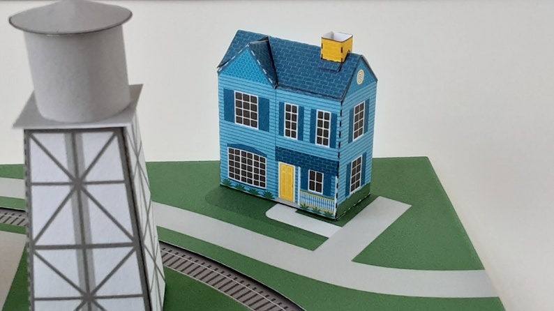 K&ouml;nnte beinhalten: Ein blaues Papiermodellhaus mit einer gelben T&uuml;r und einem wei&szlig;en Lattenzaun, neben einem grauen Papiermodell-Wasserturm.