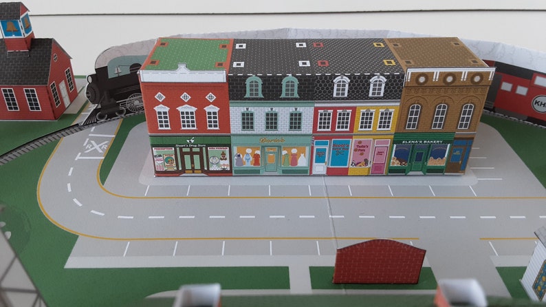 K&ouml;nnte beinhalten: Ein Miniatur-Papiermodell einer Stadt mit einem Gleis, einem roten Backsteinschulhaus und einer Reihe von bunten Ladenfronten mit gr&uuml;nen D&auml;chern. Die Ladenfronten haben Schilder mit der Aufschrift "Sweet's Toy Store", "Wardrobe", "Mother's Day" und "Elena's Bakery".
