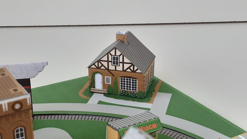 K&ouml;nnte beinhalten: Ein Papiermodell eines kleinen braunen Hauses mit einem grauen Dach und wei&szlig;en Verzierungen. Das Haus hat eine wei&szlig;e T&uuml;r, ein Fenster mit einem wei&szlig;en Rahmen und einen kleinen Schornstein. Das Haus steht auf einem gr&uuml;nen Rasen mit einem wei&szlig;en Gehweg.