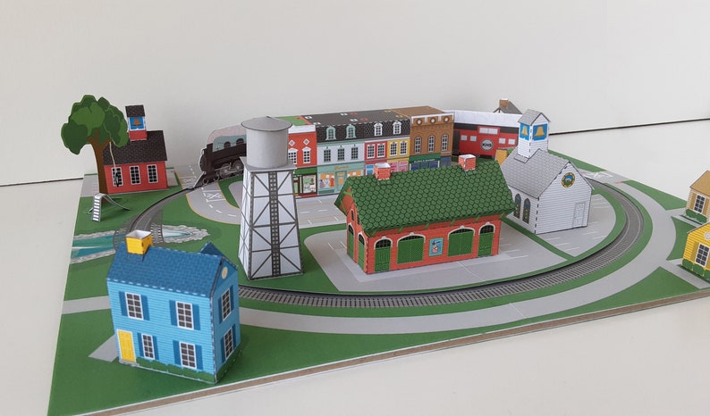 K&ouml;nnte beinhalten: Ein Papiermodell einer kleinen Stadt mit einem Gleis, einem Wasserturm, einer Kirche und mehreren H&auml;usern. Die H&auml;user sind in verschiedenen Farben gestrichen, darunter Rot, Blau, Gr&uuml;n und Gelb.
