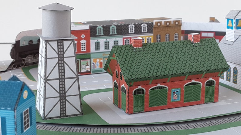 K&ouml;nnte beinhalten: Ein Papiermodell einer kleinen Stadt mit einem Wasserturm, einem Bahnhof und mehreren Geb&auml;uden. Die Geb&auml;ude sind rot, gr&uuml;n und wei&szlig; mit schwarzen Verzierungen. Der Bahnhof hat ein gr&uuml;nes Dach und rote W&auml;nde mit einem Schild, auf dem "Bahnhof" steht.