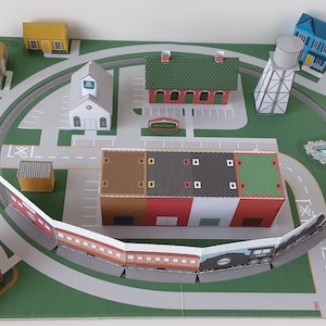 K&ouml;nnte beinhalten: Ein Papiermodell einer Stadt mit einem Gleis, einer Kirche, einem Bahnhof, einem Wasserturm, einer Fabrik und mehreren H&auml;usern. Das Gleis ist ein Kreis um die Stadt und der Zug besteht aus mehreren Wagen.