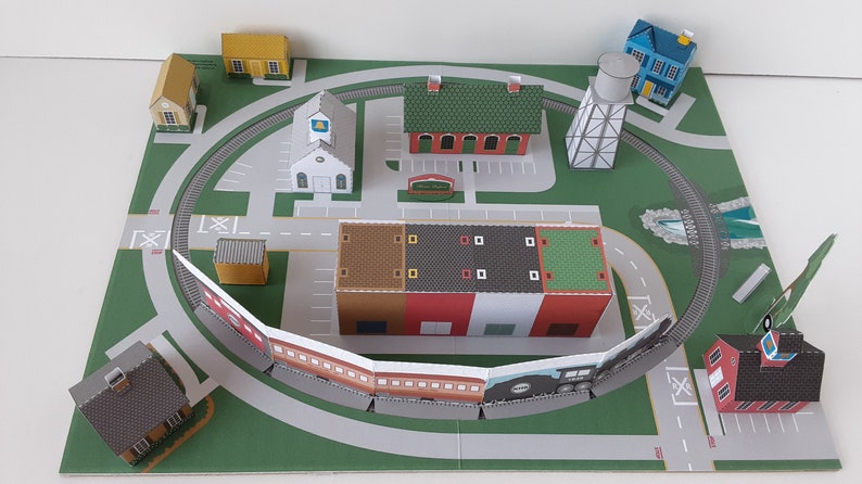 K&ouml;nnte beinhalten: Ein Papiermodell einer Stadt mit einem Gleis, einer Kirche, einem Bahnhof, einem Wasserturm, einer Fabrik und mehreren H&auml;usern. Das Gleis ist ein Kreis um die Stadt und der Zug besteht aus mehreren Wagen.