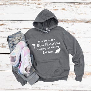 Könnte beinhalten: Ein grauer Kapuzenpullover mit dem Text "All I want to do is Drink Margaritas And hang out with my Chickens." Der Hoodie wird mit einer blauen Jeans und einem Paar weiß-rosa Turnschuhen kombiniert.