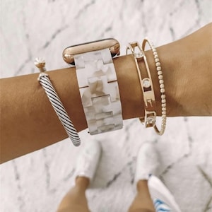 Op de afbeelding: Een goudkleurige smartwatch met een wit en grijs gemarmerde hars schakelband, vergezeld door een zilverkleurige gedraaide kabelarmband, een goudkleurige manchetarmband met drie ronde heldere stenen en een dunne goudkleurige kralenkettingarmband.