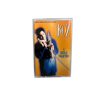 Könnte beinhalten: Eine Musikkassette mit Dave Koz's "A Sensual Collection". Das Cover zeigt einen Mann in einem blauen Anzug, der ein Saxophon und ein Telefon hält. Der Hintergrund ist orange mit dem Text "KOZ" in großen Buchstaben.