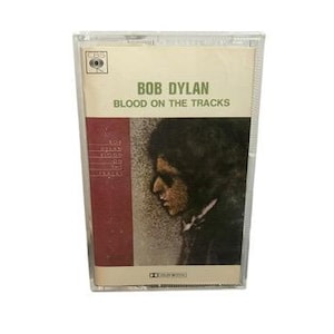 May include: A vintage cassette tape of Bob Dylan's album "Blood on the Tracks." The cassette case is clear plastic, with a cream-colored label featuring the album title and artist's name in green text. The cover art shows a profile of a person.