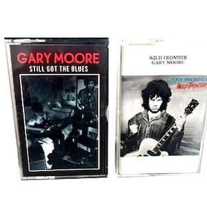 以下が含まれることがあります： 2つのヴィンテージカセットテープ。1つ目は「GARY MOORE STILL GOT THE BLUES」の文字が赤と白で表示されています。2つ目のテープには「WILD FRONTIER GARY MOORE」と書かれており、ギターを弾く男性の写真が写っています。