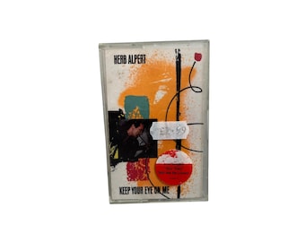 May include: A cassette tape of Herb Alpert's album "Keep Your Eye on Me." The cover art includes orange and teal paint splatters, a black line, and a photo of a man. The album title is printed at the bottom of the cassette case.