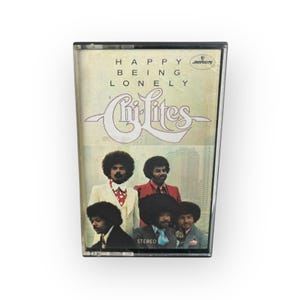 以下が含まれることがあります： The Chi-Litesの「Happy Being Lonely」のアルバムカバーが印刷された、ヴィンテージのカセットテープ。カバーアートには、アフロヘアのスーツ姿の男性4人が写っています。アルバムタイトルとバンド名がカバーに印刷されています。