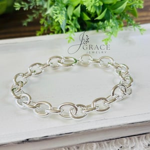 May include: A polished silver chain link bracelet featuring a series of interconnected oval-shaped metal loops. The jewelry piece is displayed with the logo J GRACE JEWELRY printed in black text above the center of the bracelet.