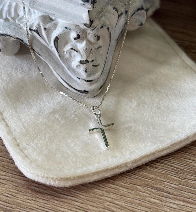 May include: A silver-toned necklace featuring a simple, polished cross pendant suspended from a delicate box chain. The cross has clean, straight lines and a smooth, reflective surface, showcasing a classic design that hangs from a small jump ring.