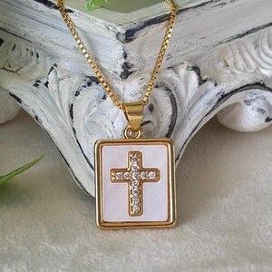 May include: Gold-toned necklace featuring a square pendant with a white background and a cross design. The cross is adorned with small, sparkling crystals. The necklace is a religious accessory.