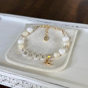 May include: A beaded bracelet with the word "HOPE" spelled out in white letter beads. The bracelet features white and cream-colored beads, clear crystal accents, and a gold-colored bird charm. The bracelet is displayed on a white fabric.