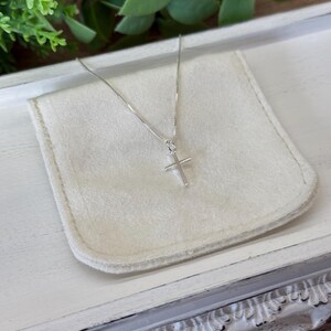May include: A delicate silver-toned necklace featuring a simple, slender cross pendant suspended from a fine box chain. The metallic cross has a smooth, polished finish and hangs centered, showcasing a minimalist design with clean, straight lines.