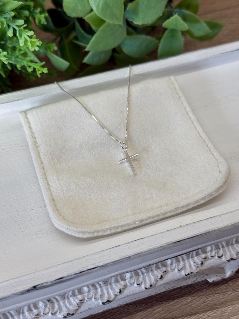 May include: A delicate silver-toned necklace featuring a simple, slender cross pendant suspended from a fine box chain. The metallic cross has a smooth, polished finish and hangs centered, showcasing a minimalist design with clean, straight lines.