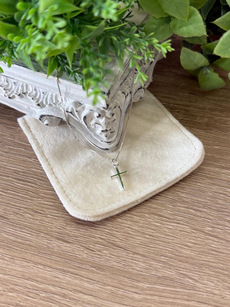 May include: A delicate silver-toned necklace featuring a simple, polished cross pendant suspended from a thin box chain. The metal surface reflects light, highlighting the clean lines and minimalist design of the cross and the fine link chain structure.